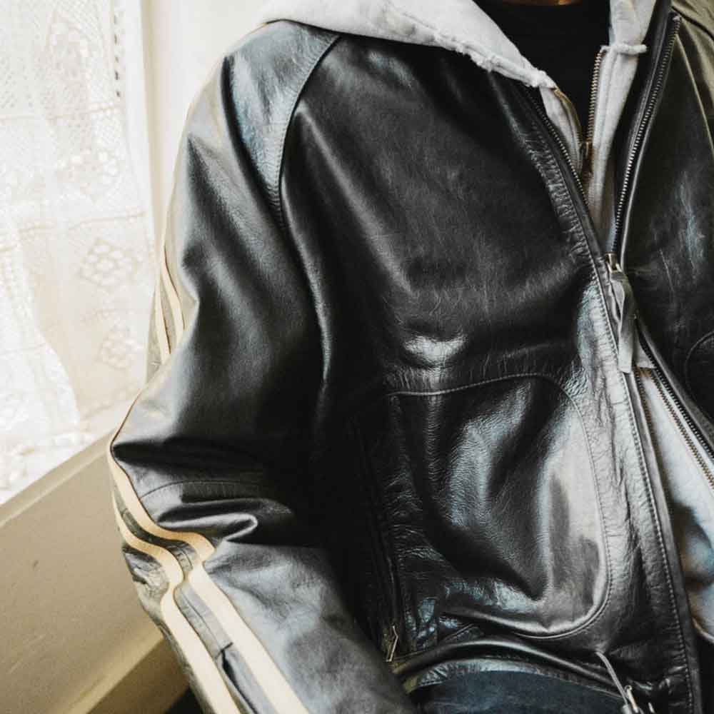A close-up of a person wearing a black leather jacket with a light gray hood and beige stripes on the sleeves, photographed indoors near a window.
