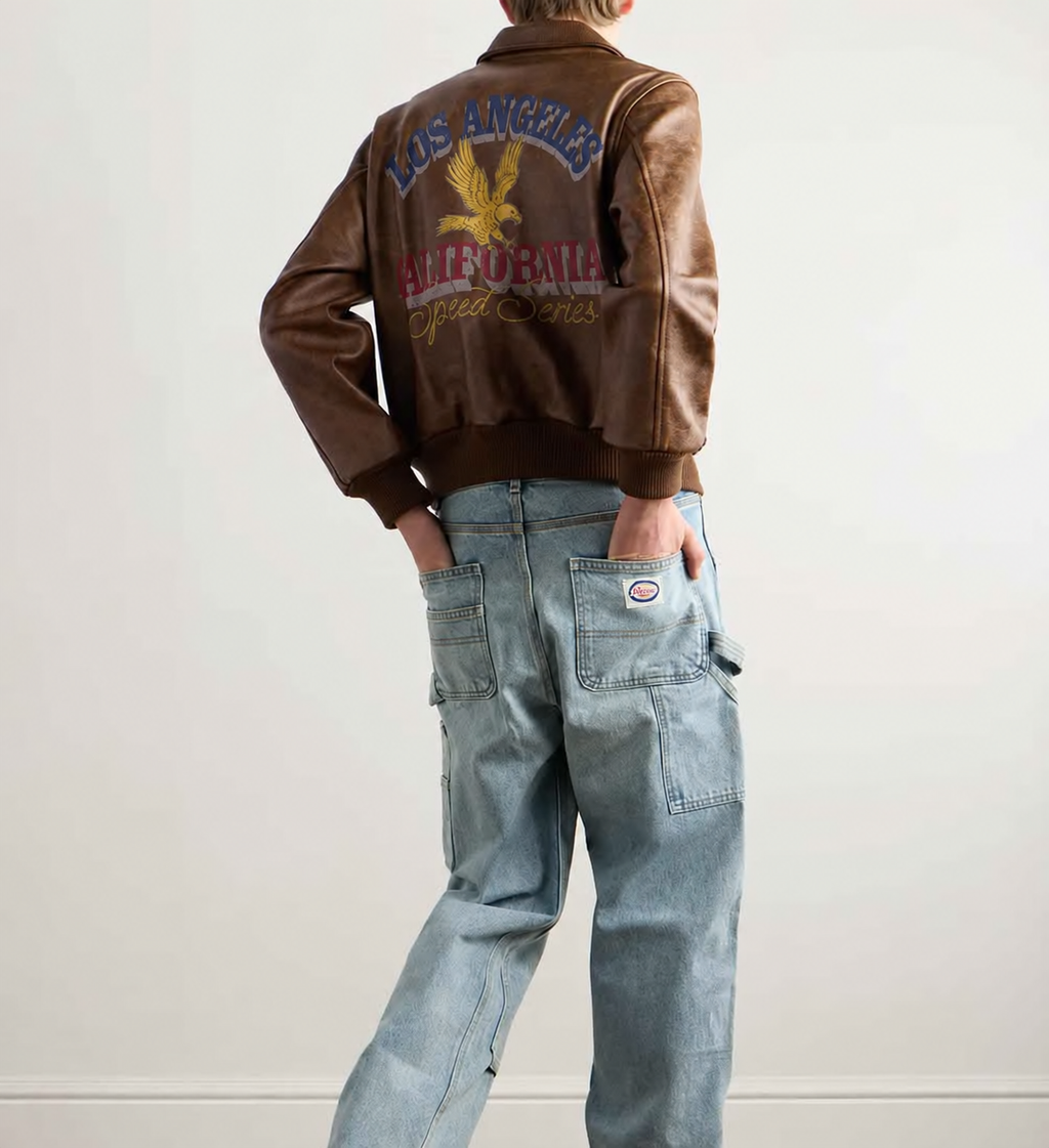 A person shown from the back wearing a brown leather bomber jacket with a large embroidered Los Angeles California graphic on the back, paired with light blue jeans.