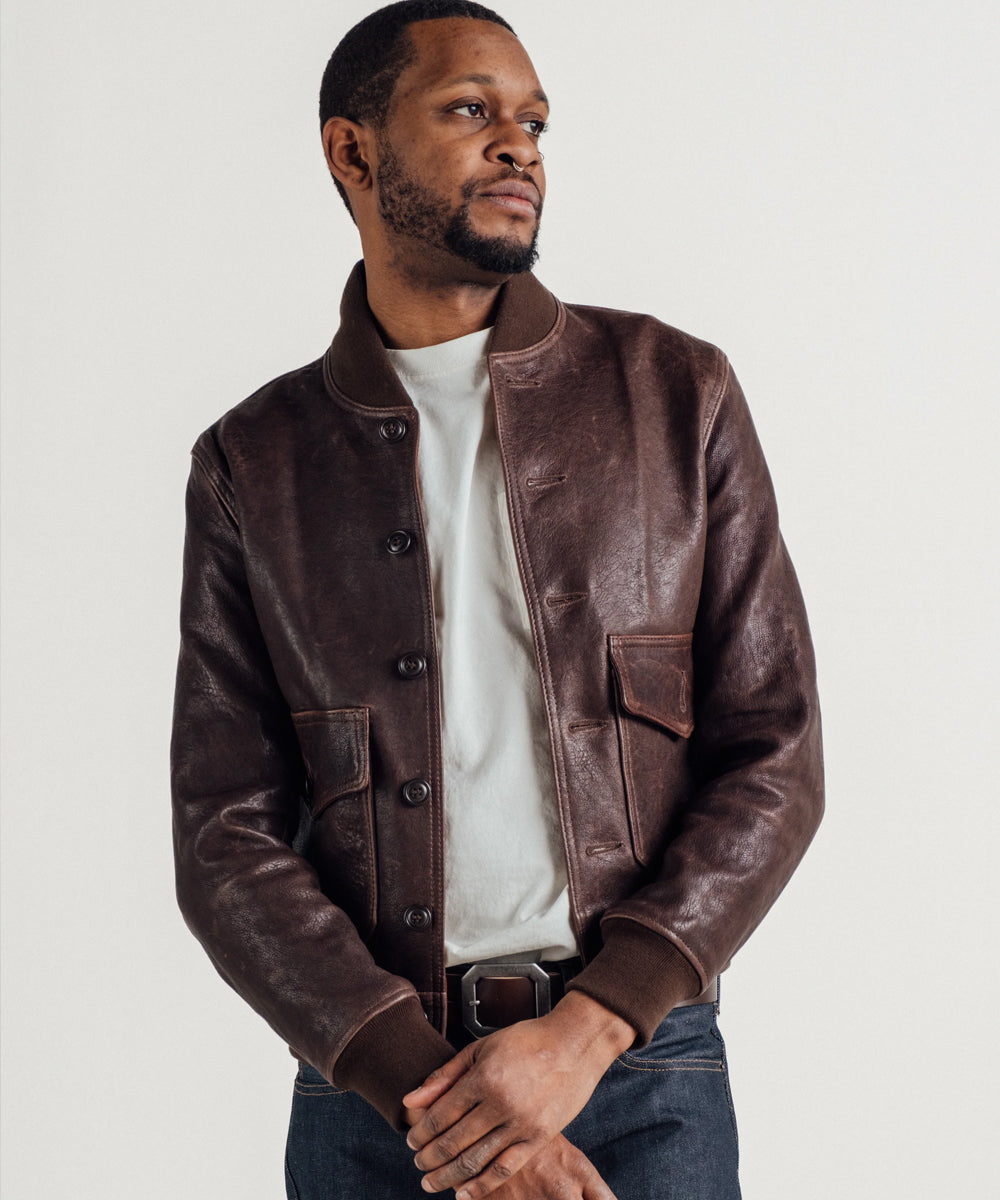 A man wearing a dark brown leather bomber jacket over a white t-shirt and blue jeans, standing against a light gray studio background.