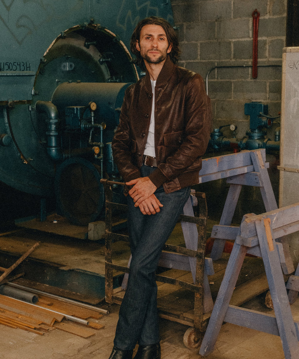 A man wearing a brown leather jacket, white t-shirt, and dark blue jeans stands in a workshop setting with a large blue machine behind him.