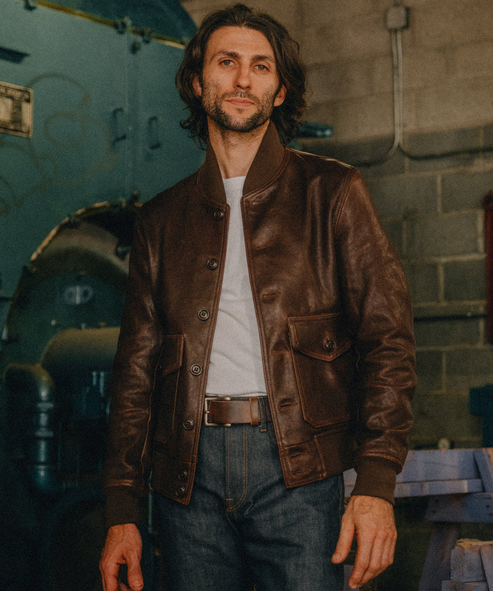 A man wearing a brown leather bomber jacket over a white t-shirt and blue jeans, standing in an industrial workshop setting.