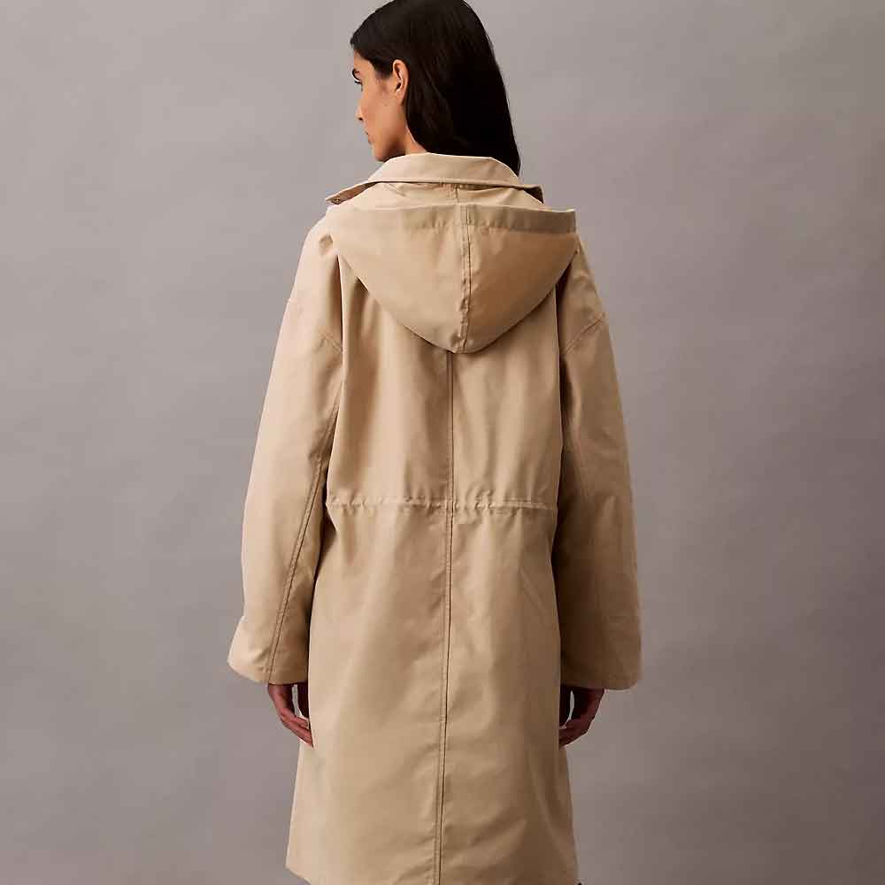 A woman shown from the back wearing a long hooded coat in a light tan or beige color, set against a plain gray studio background.