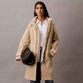 A woman modeling a mid-length beige Anorak Rain Jacket over a white button-up shirt and dark jeans, holding a black shoulder bag against a plain gray studio background.