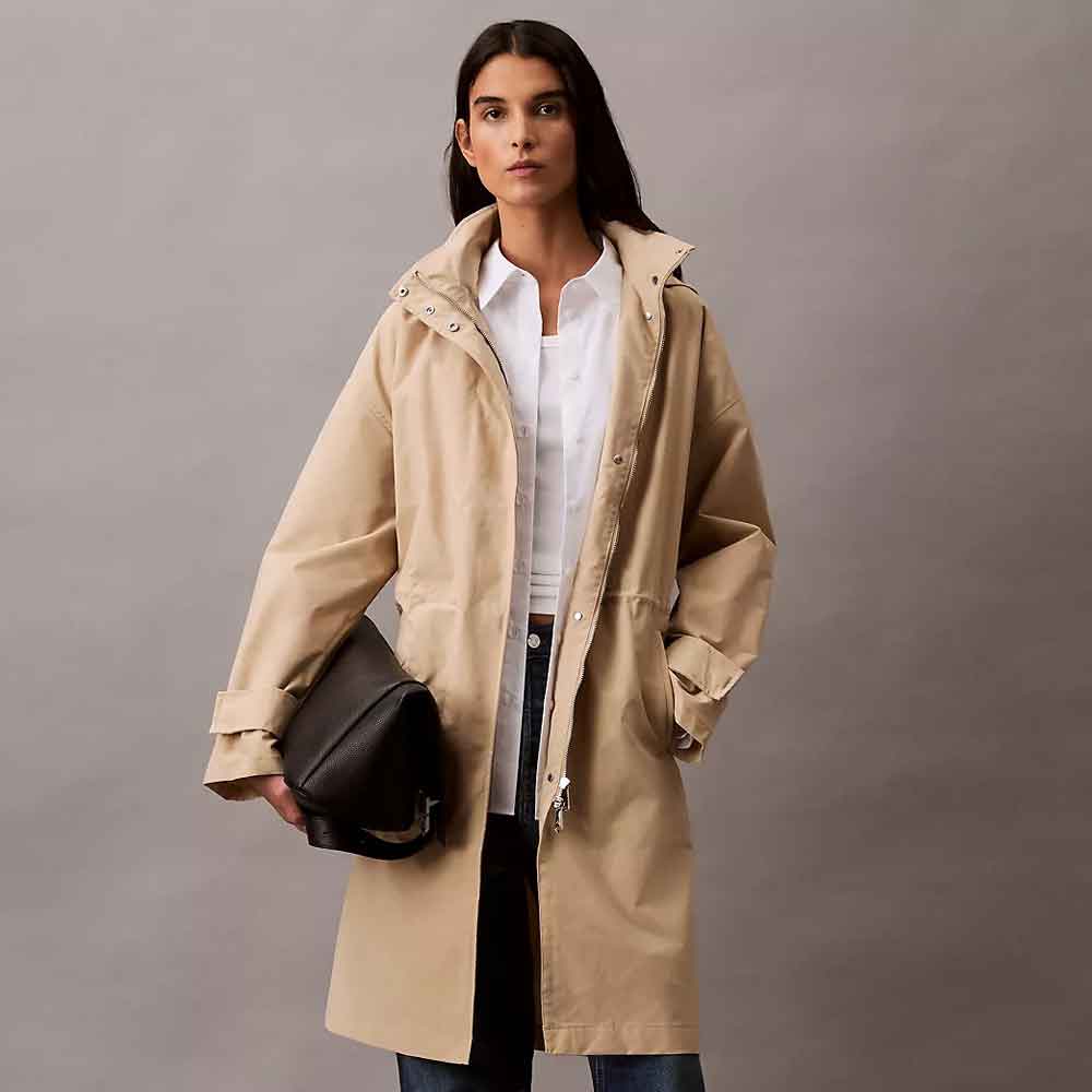 A woman modeling a mid-length beige Anorak Rain Jacket over a white button-up shirt and dark jeans, holding a black shoulder bag against a plain gray studio background.