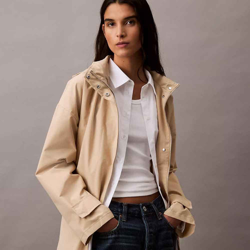 A woman wearing a beige hooded Anorak Rain Jacket over a white collared shirt and dark blue jeans, posed against a neutral gray studio background.