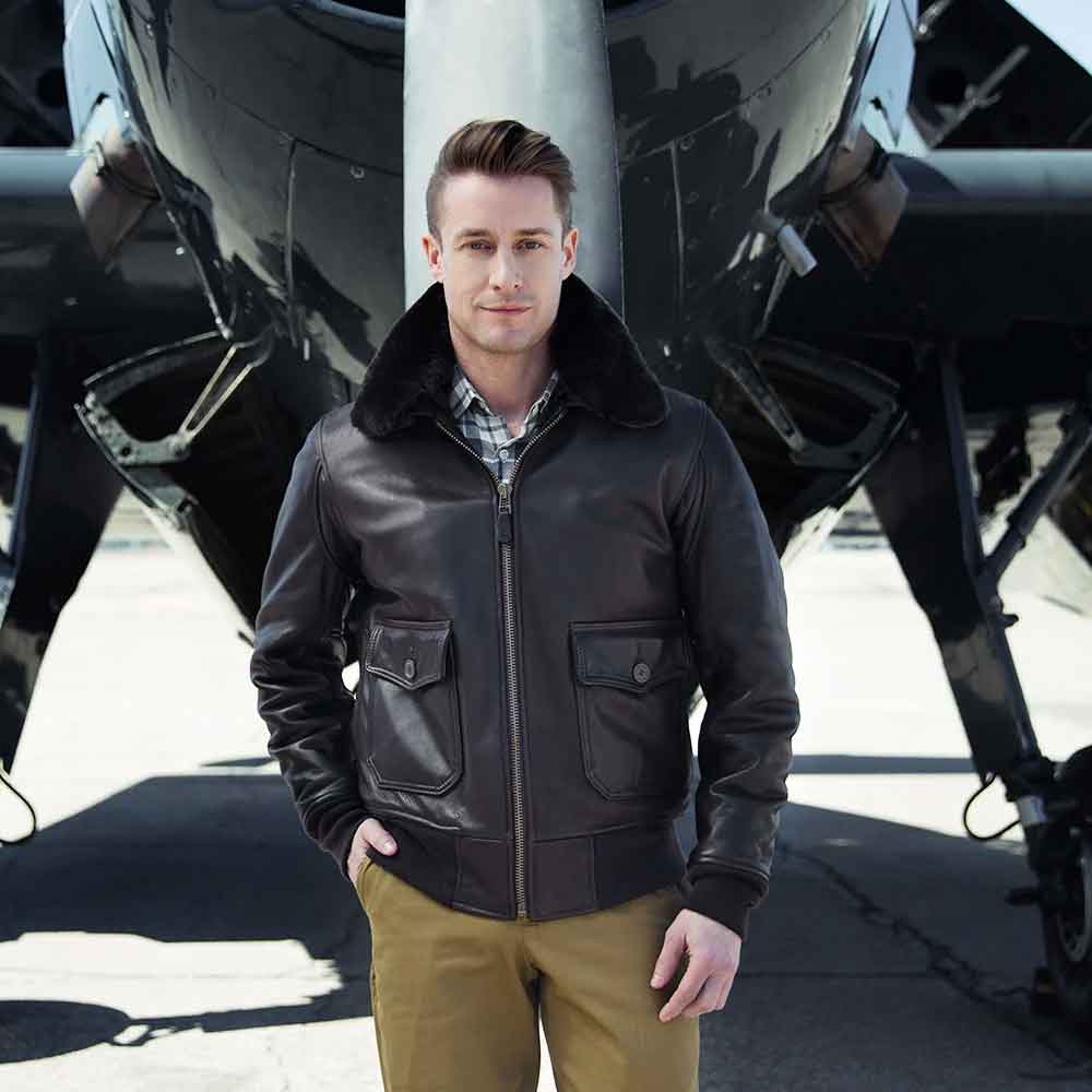 A man wearing a dark brown leather aviator jacket with a black shearling collar, standing in front of a black airplane on an airfield.