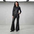 A woman modeling a black leather jumpsuit with long sleeves, a matching belt, and flared legs, standing in a studio with a light gray background.