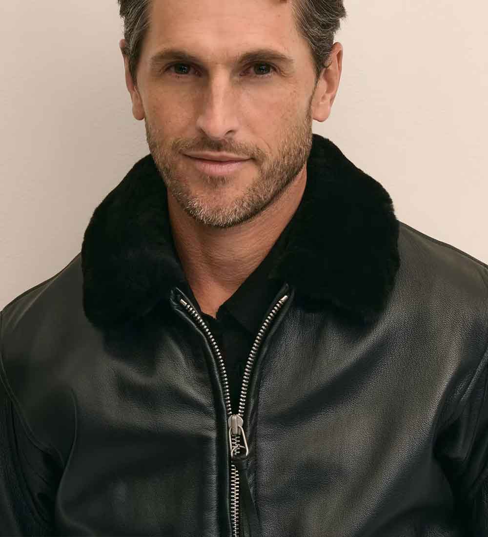A close-up of a man wearing a black leather jacket with a thick black fur collar, photographed against a plain light beige background.