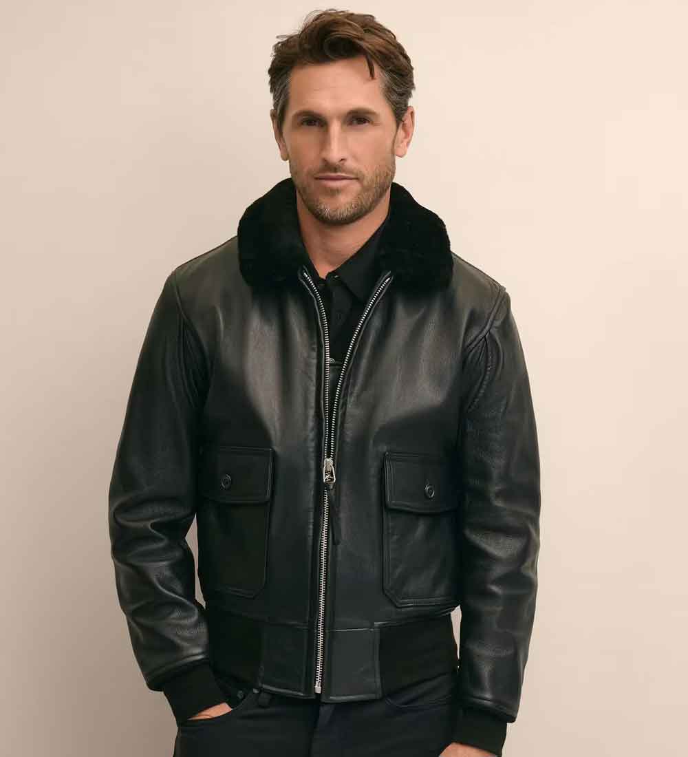 A man wearing a black leather bomber jacket with a black fur collar, standing against a plain beige background.