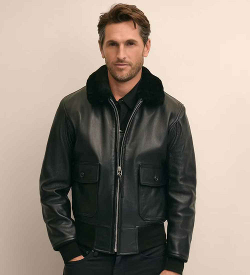 A man wearing a black leather bomber jacket with a black fur collar, standing against a plain beige background.