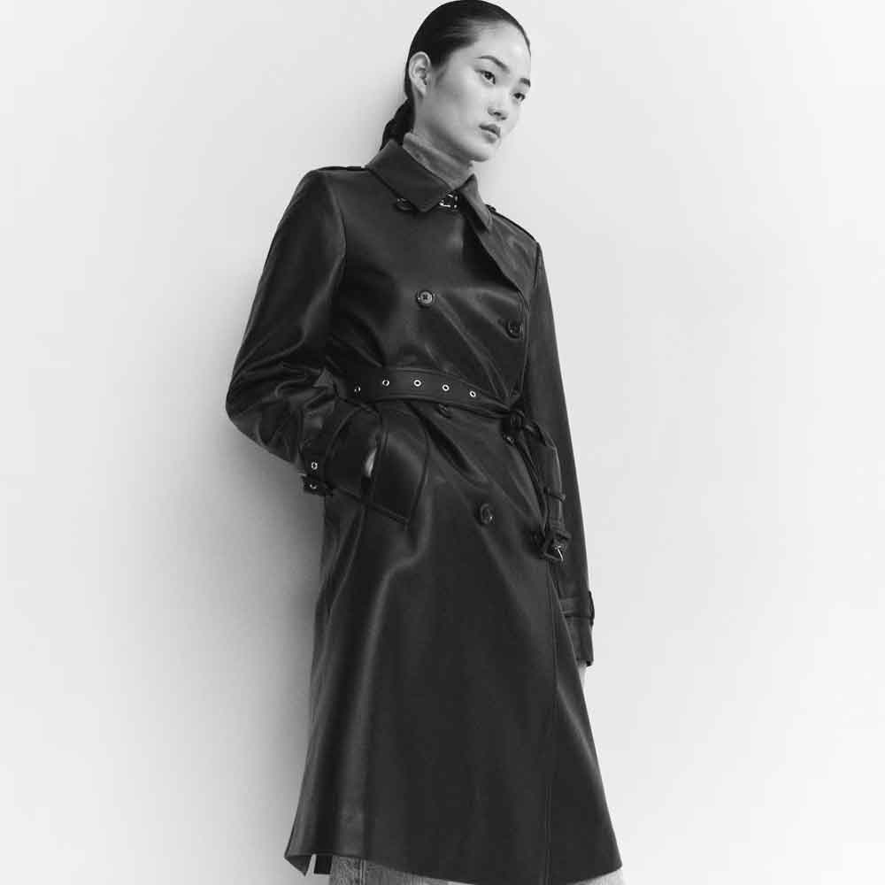 A model wearing a black leather trench coat with a matching tie belt, photographed in black and white against a plain light background.
