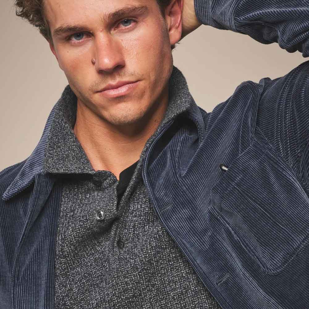 A close-up of a man wearing a dark navy corduroy overshirt over a textured gray knit polo shirt, set against a plain beige studio background.