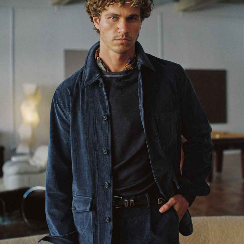 A man wearing a dark navy corduroy overshirt over a black shirt and trousers, standing in a softly lit indoor room.