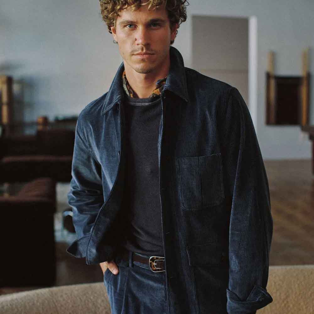 A man wearing a dark navy blue corduroy jacket over a dark crewneck sweater, paired with matching navy corduroy trousers and a brown belt, standing in a softly lit indoor setting.