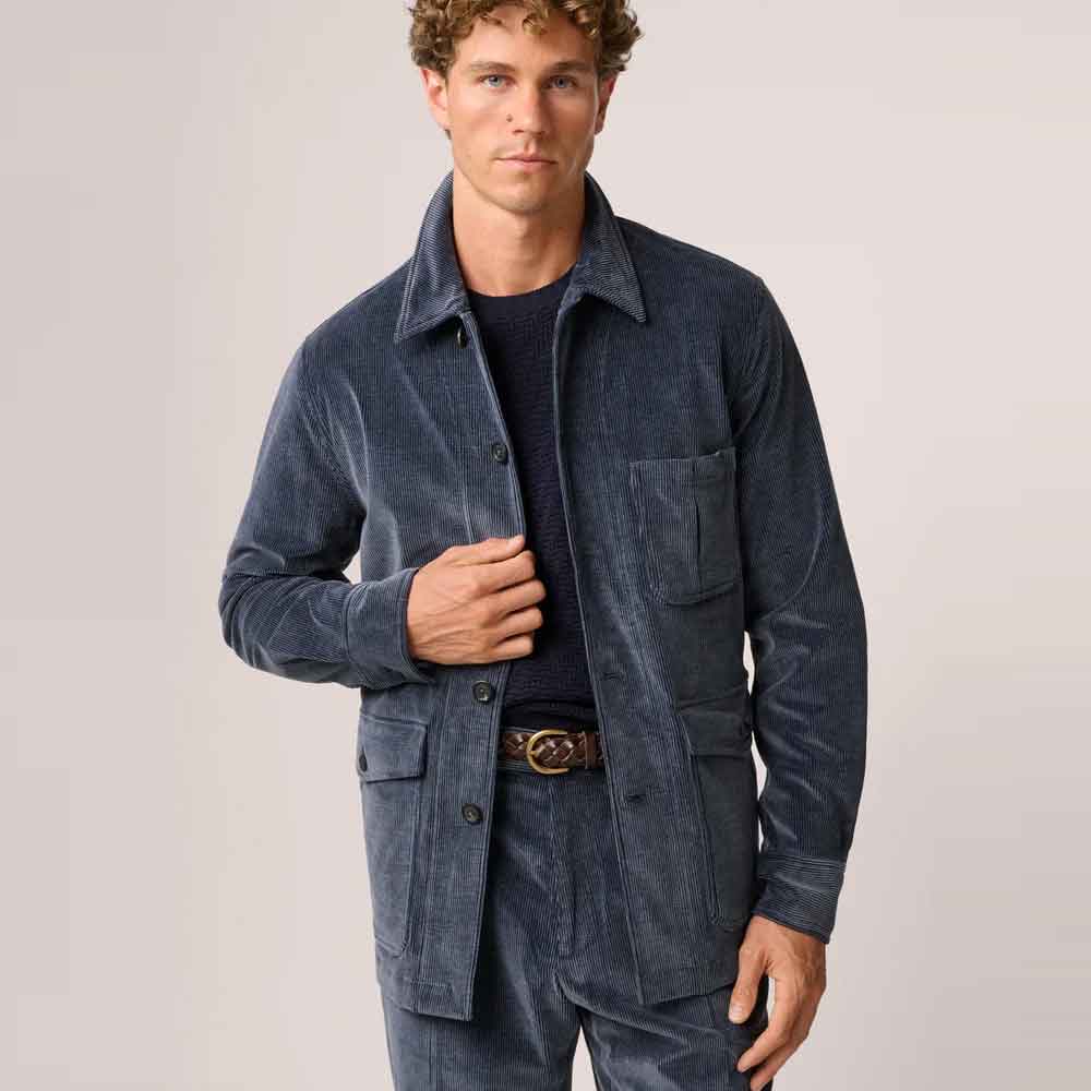 A man wearing a dark blue corduroy utility jacket over a dark navy shirt, paired with matching corduroy trousers and a brown belt, against a light gray studio background.