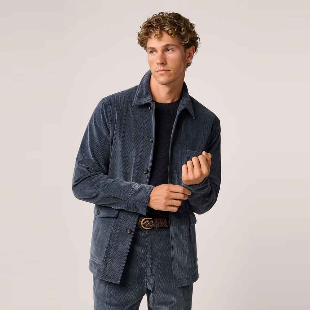 A man wearing a dark Blue Corduroy Jacket and matching corduroy trousers over a black t-shirt and brown belt, standing against a light gray studio background.