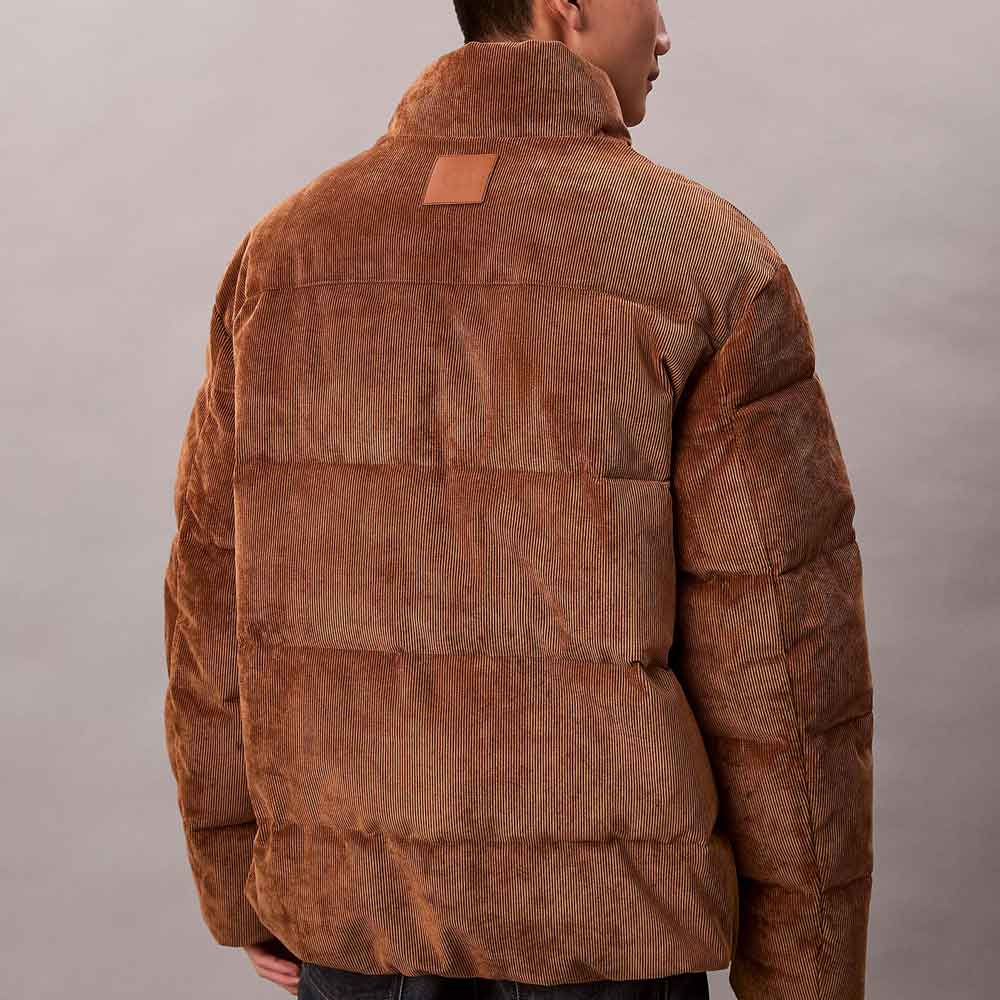 A man viewed from the back wearing a brown puffer jacket with a textured corduroy-like finish and a small rectangular patch on the upper back, against a plain light gray studio background.