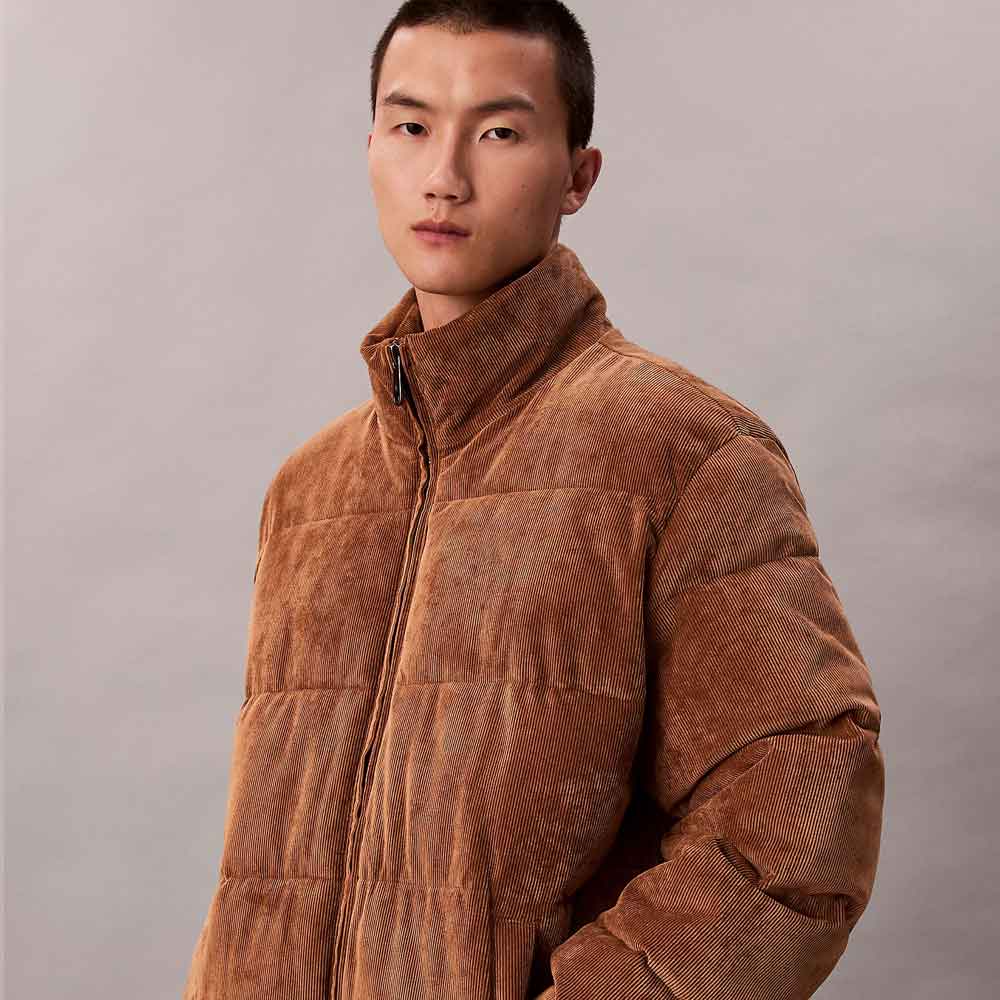A man wearing a brown corduroy puffer jacket with a high stand-up collar, photographed against a neutral gray studio background.