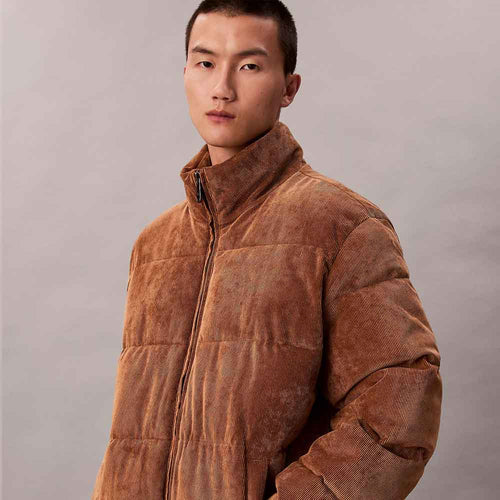 A man wearing a brown corduroy puffer jacket with a high stand-up collar, photographed against a neutral gray studio background.