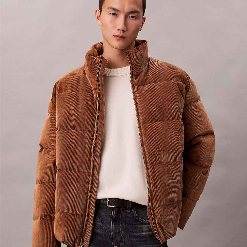 A man wearing a brown puffer jacket with a high collar and zippered front, layered over a cream t-shirt and dark jeans, against a plain gray studio background.