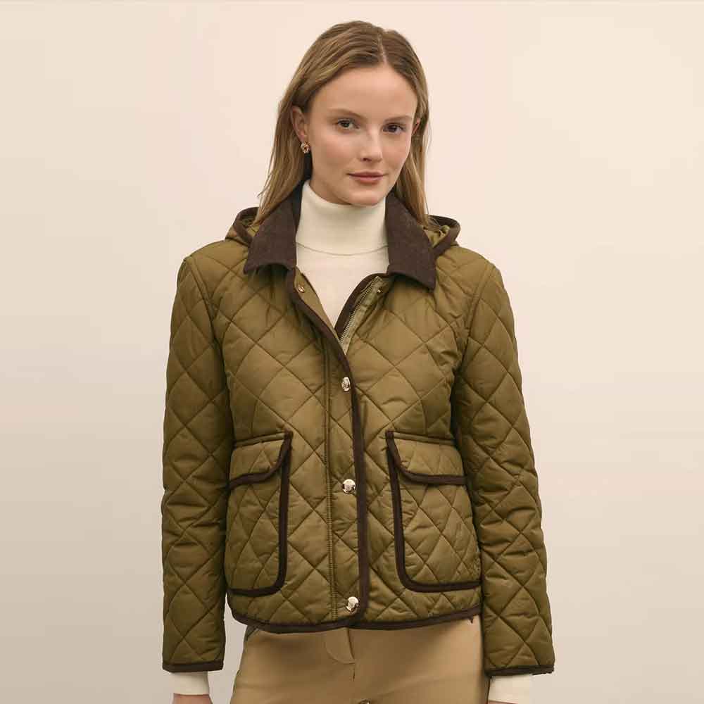 A woman modeling a quilted olive green jacket with dark brown trim and a matching hood, worn over a white turtleneck sweater and beige pants, against a plain light beige background.