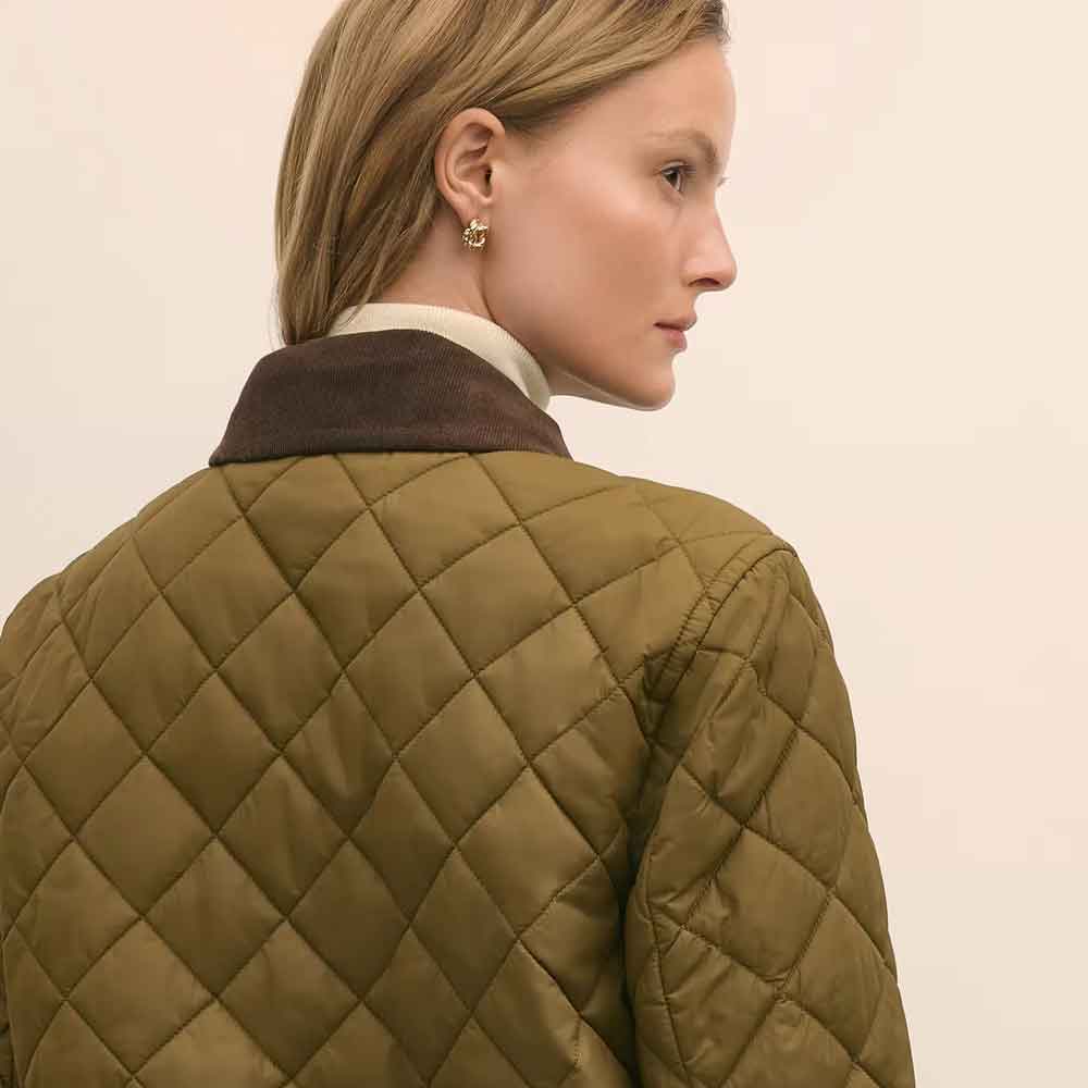 A close-up side profile of a woman wearing a cropped quilted jacket in olive green with a dark brown collar, set against a plain light beige background.