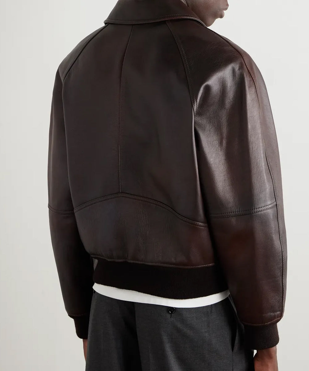 A dark brown leather jacket with a high collar, shown from the back on a model. The jacket features a ribbed black hem and cuffs, and is styled over a white t-shirt and dark gray trousers against a plain light gray background.