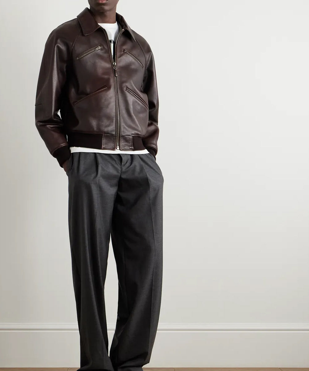 A model wearing a dark brown leather bomber jacket, black wide-leg trousers, and a white undershirt, standing against a plain off-white wall.