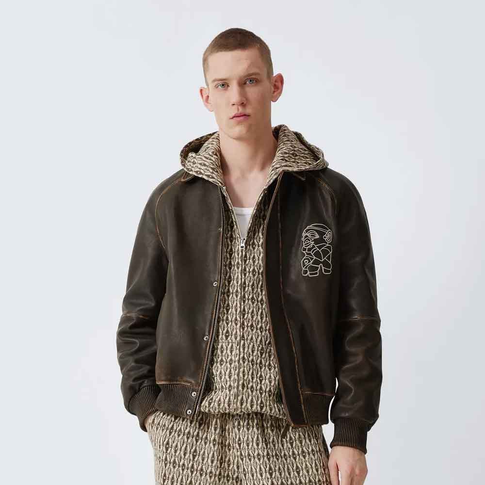 A male model wearing a dark brown leather bomber jacket with a patterned knit hoodie underneath, set against a plain light gray studio background.