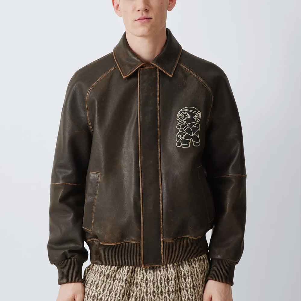 A model wearing a dark brown leather bomber jacket with a white embroidered cartoon bear on the left chest, paired with a patterned knit skirt, against a light gray studio background.