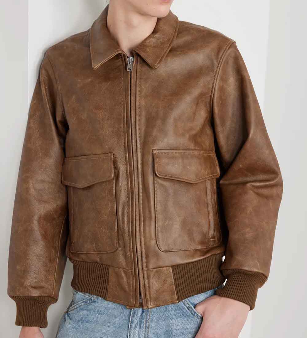 A person wearing a brown leather bomber jacket with a front zipper, two large flap pockets, and ribbed knit collar, cuffs, and hem, paired with light blue jeans against a plain light background.