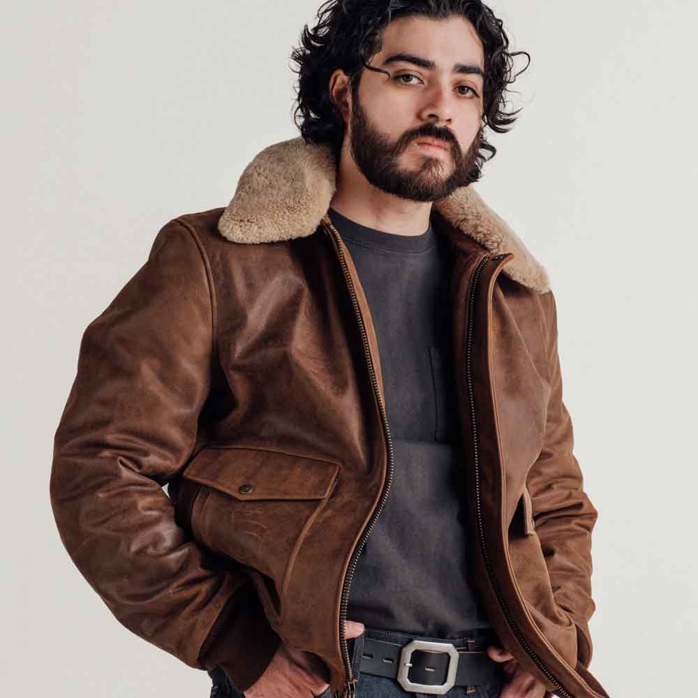 A man wearing a brown leather aviator-style jacket with a thick beige shearling collar, layered over a dark gray t-shirt and a black belt, photographed against a plain light gray studio background.
