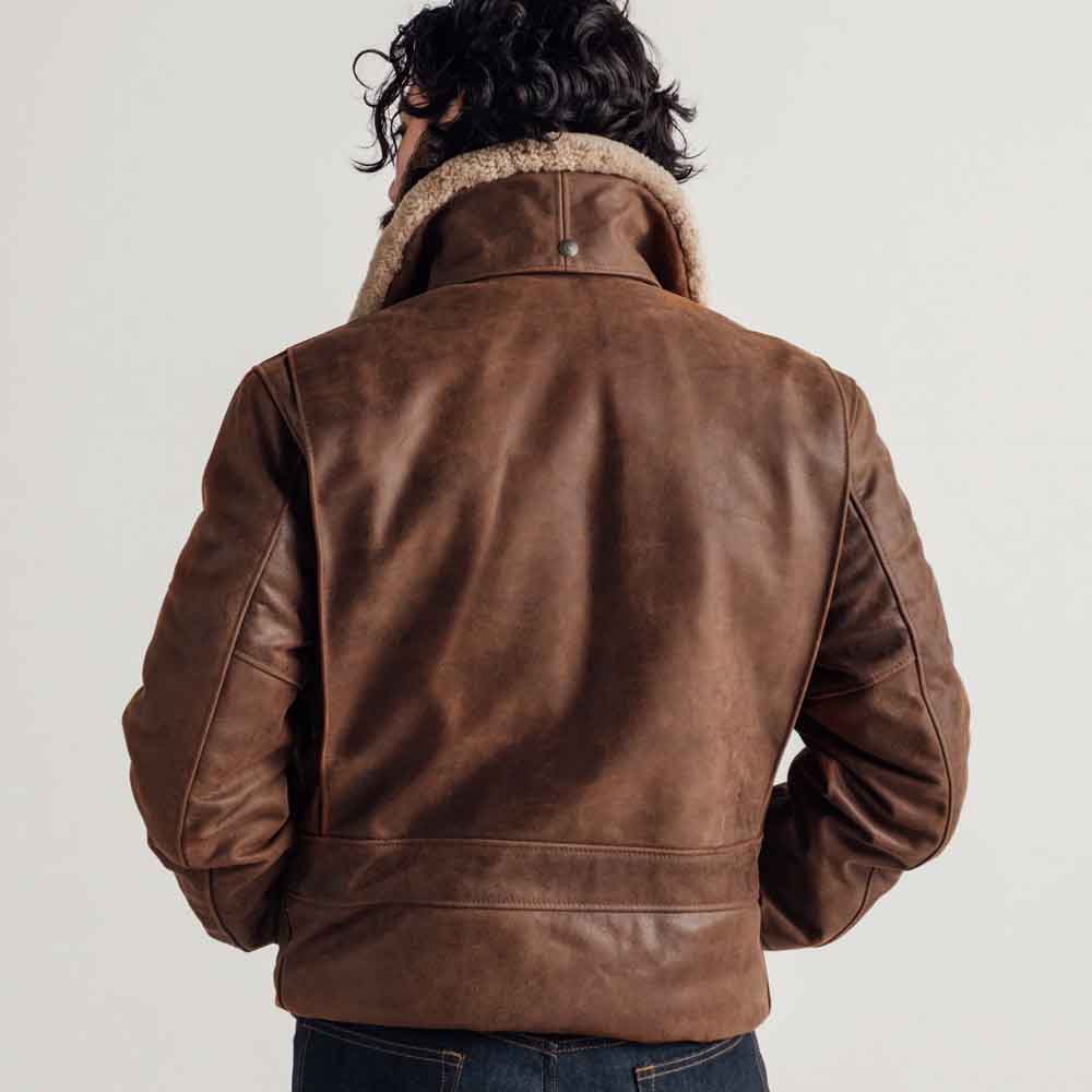 A man viewed from behind wearing a brown leather aviator-style jacket with a beige shearling collar, paired with dark blue jeans, against a plain light gray studio background.