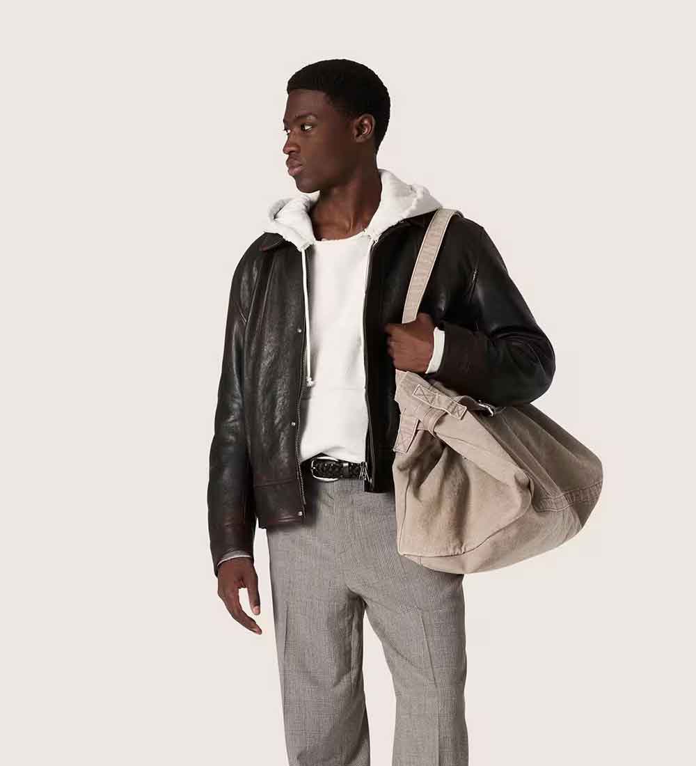 A man wearing a black leather jacket over a white hoodie, gray trousers, and a black belt, carrying a large beige shoulder bag against a light off-white background.