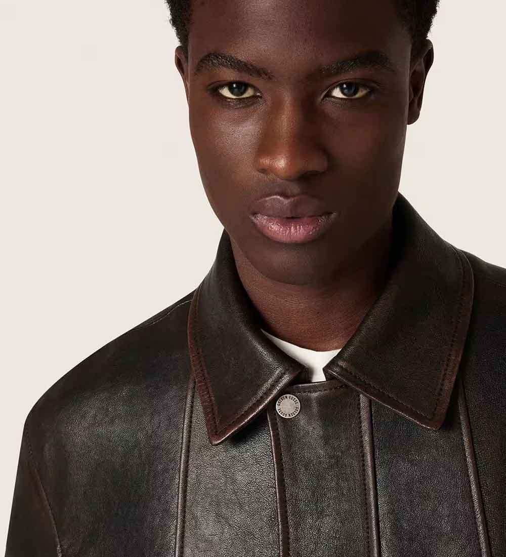 A close-up portrait of a man wearing a dark brown leather jacket with a pointed collar and a silver snap button, set against a solid light beige background.