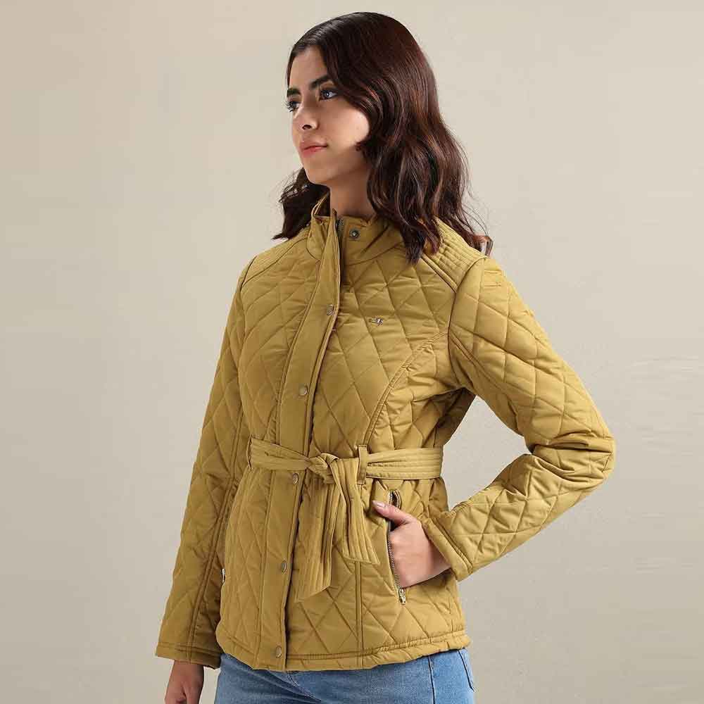 A woman wearing a quilted mustard yellow jacket with a matching waist tie belt, paired with blue jeans, set against a plain beige studio background.