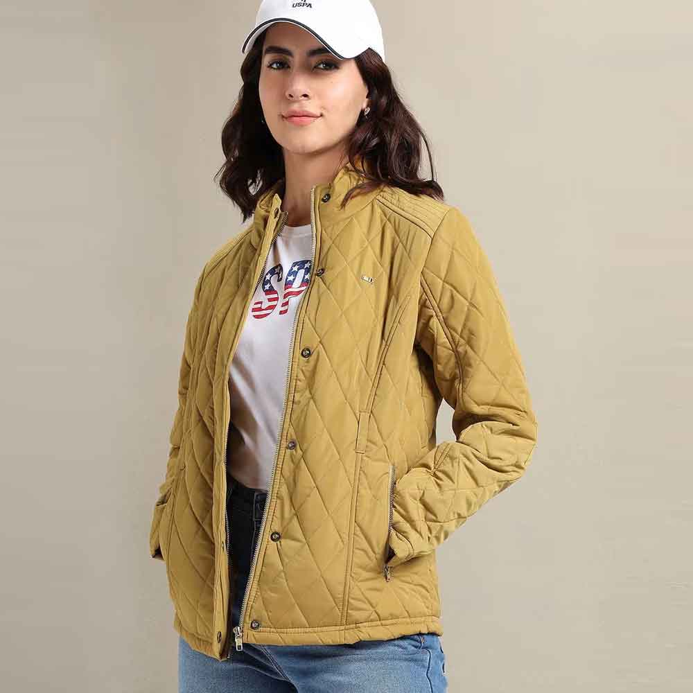 A woman wearing a quilted mustard yellow jacket over a white graphic t-shirt and blue jeans, paired with a white baseball cap, set against a plain beige studio background.