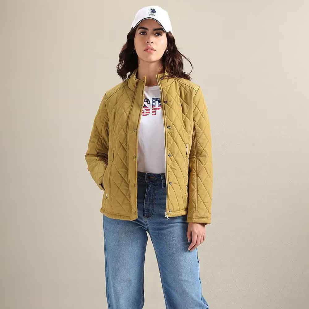 A woman modeling a quilted mustard yellow jacket over a white graphic t-shirt and blue jeans, wearing a white baseball cap, against a plain beige studio background.
