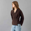 A woman wearing a dark brown Quilted Kimono Jacket for Women in Cotton with a tie waist, paired with light blue jeans, standing against a solid gray studio background.