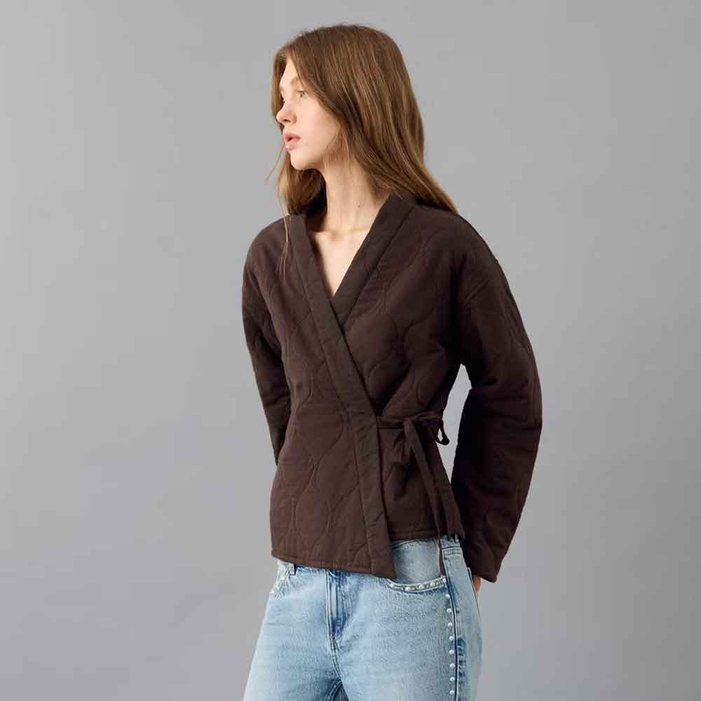 A woman wearing a dark brown Quilted Kimono Jacket for Women in Cotton with a tie waist, paired with light blue jeans, standing against a solid gray studio background.