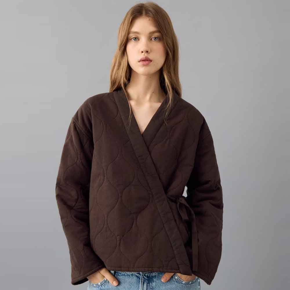 A woman modeling a dark brown quilted wrap jacket with a tie waist, paired with blue jeans, against a solid gray studio background.