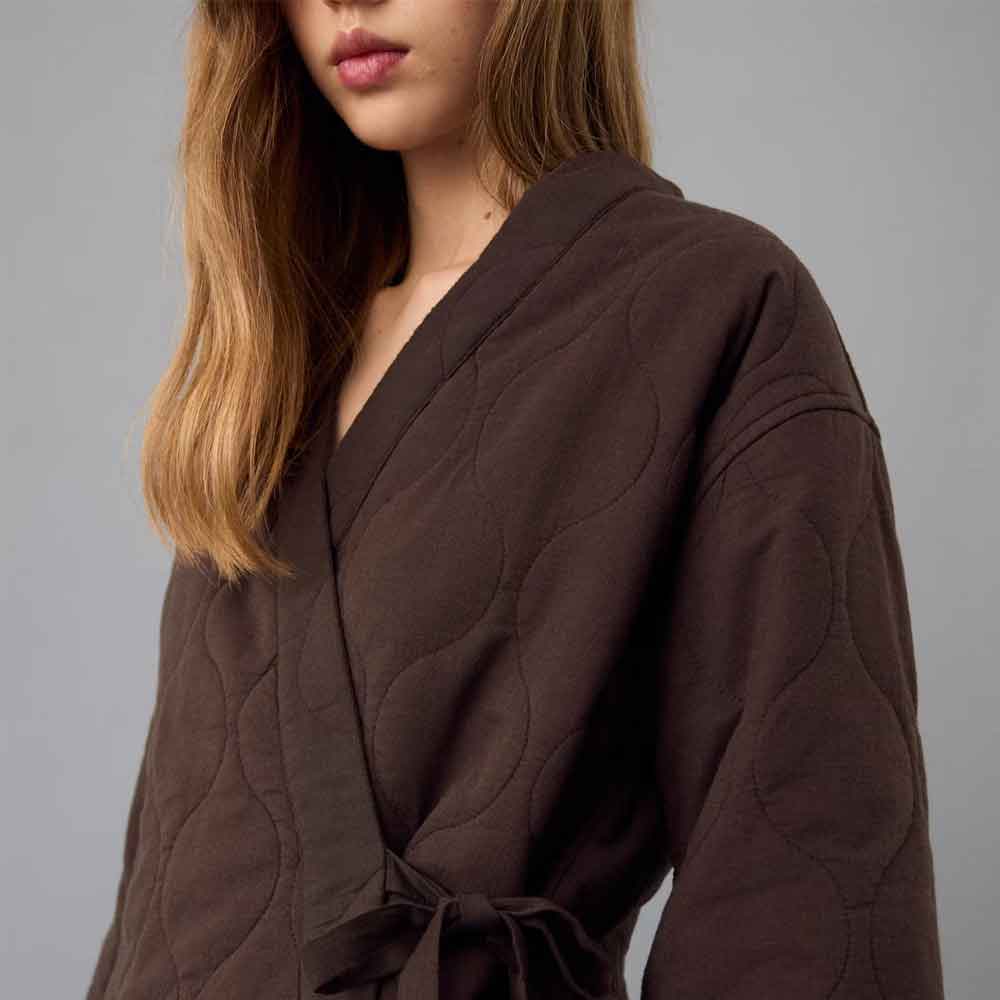 A close-up of a woman wearing a dark brown Quilted Kimono Jacket with a tie waist, shown against a plain gray studio background.
