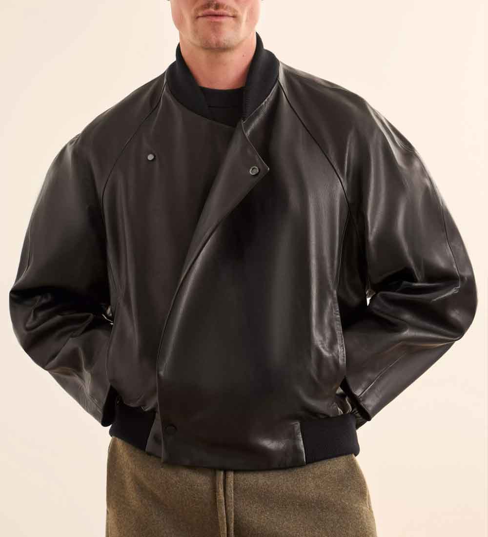 A man wearing a black leather bomber jacket with a high ribbed collar, asymmetrical snap closure, and elasticized hem and cuffs, paired with olive brown trousers against a light beige background.
