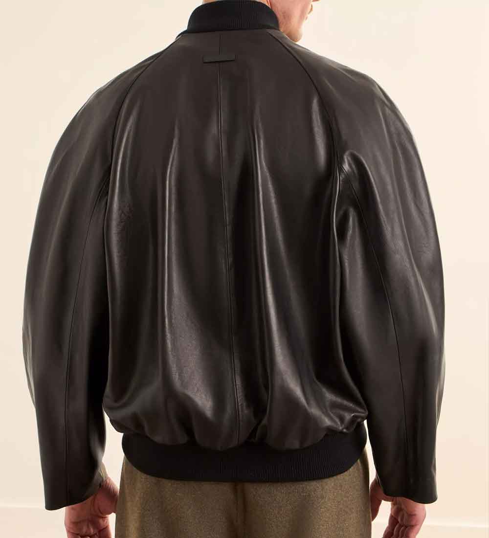 Rear view of a person wearing a black leather bomber jacket with black ribbed collar, cuffs, and hem, paired with olive green trousers, against a plain off-white background.