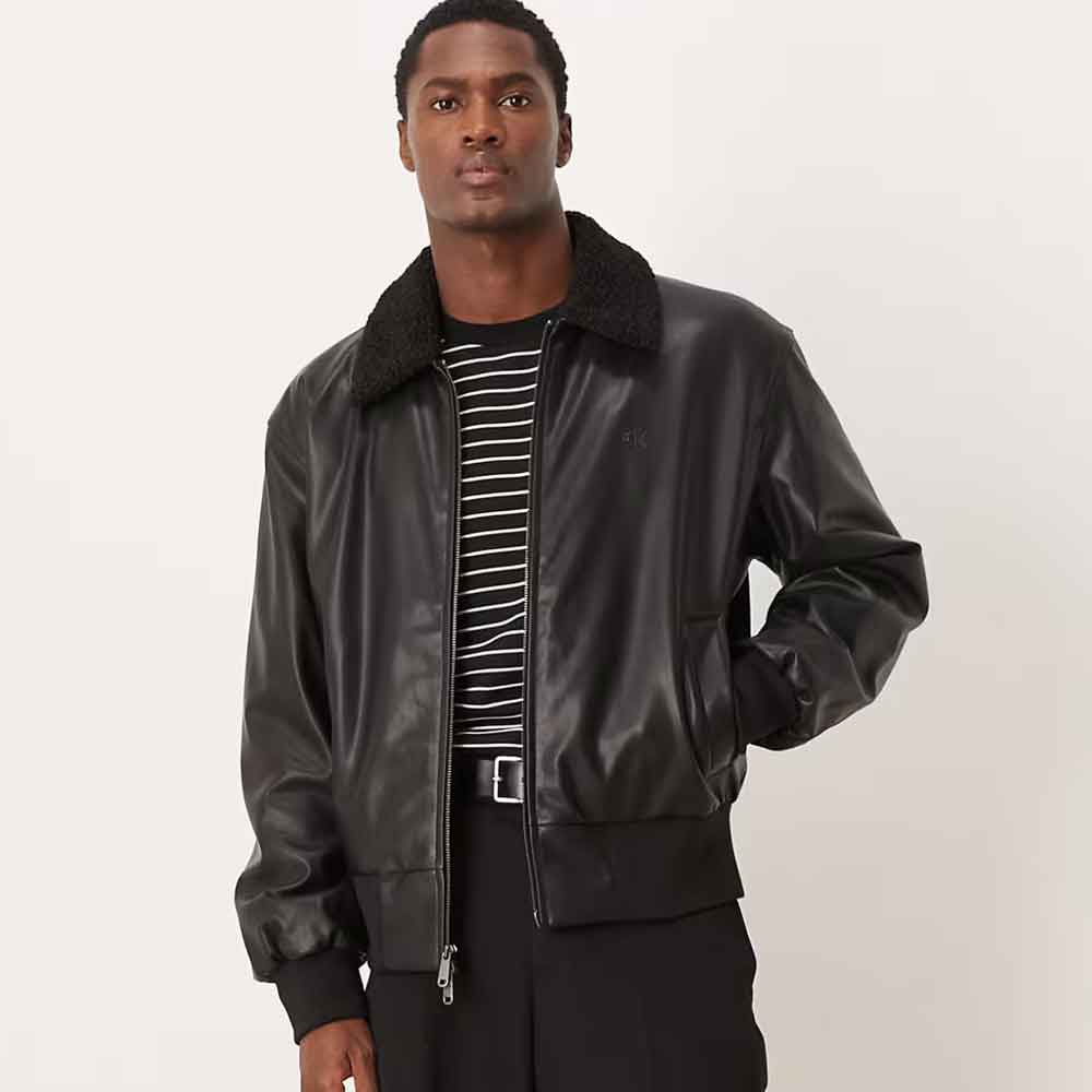 A man wearing a black faux leather bomber jacket with a dark sherpa collar, layered over a black and white striped shirt and black trousers, photographed against a plain light gray studio background.