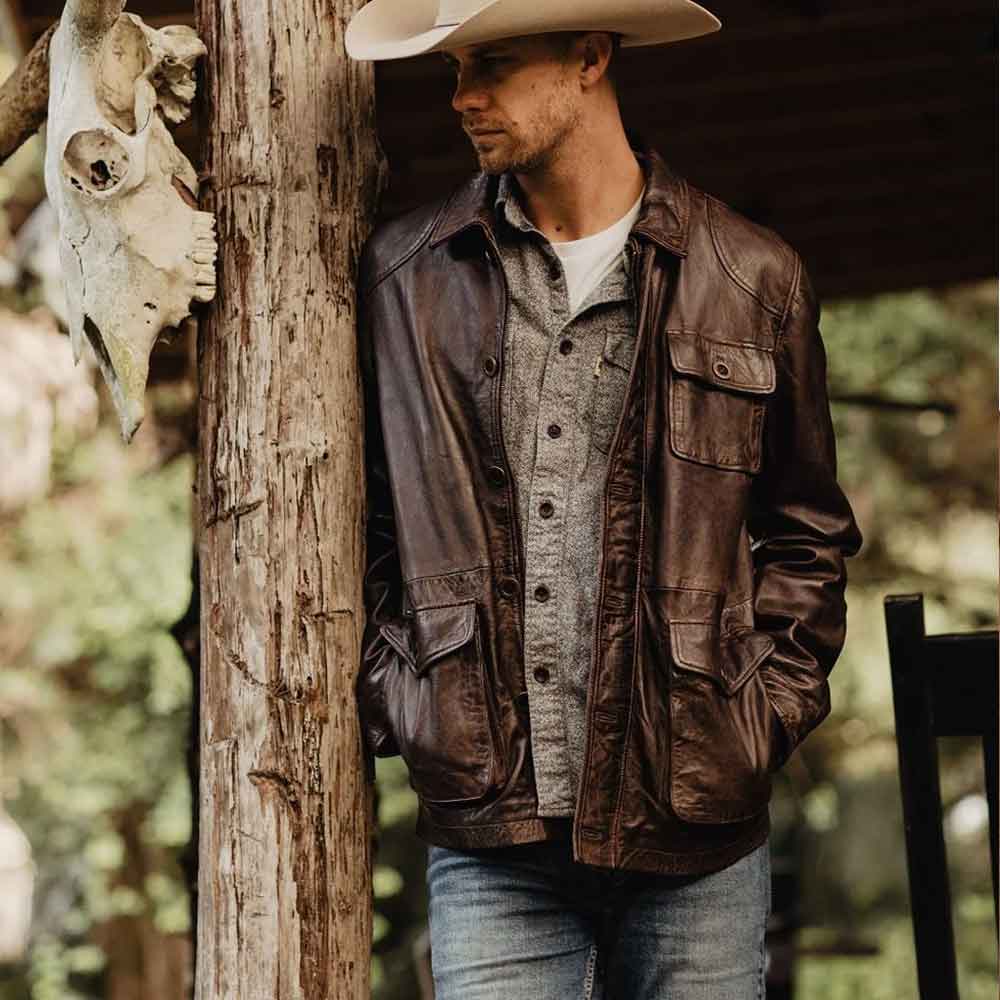 Man wearing a cowboy hat and Vintage Barn Jacket leaning against a wooden post with a blurred natural background