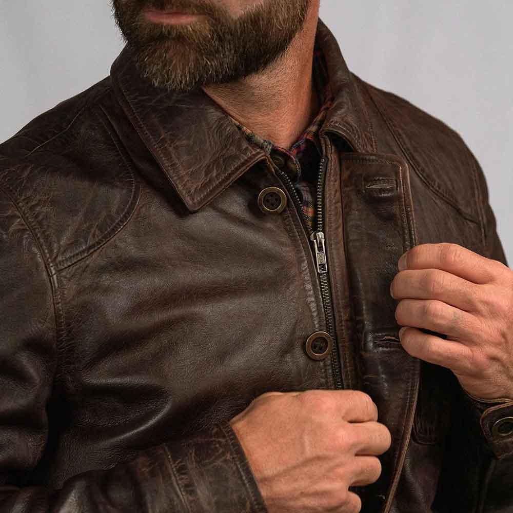 A close-up of a man wearing a dark brown leather jacket, adjusting the front zipper with one hand while pulling the collar with the other.
