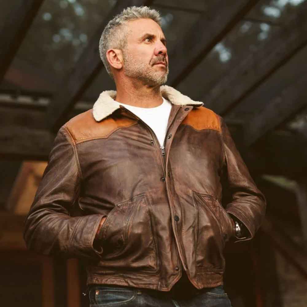 A man wearing a brown leather jacket with a cream faux shearling collar and tan shoulder panels, standing in a dimly lit rustic interior.
