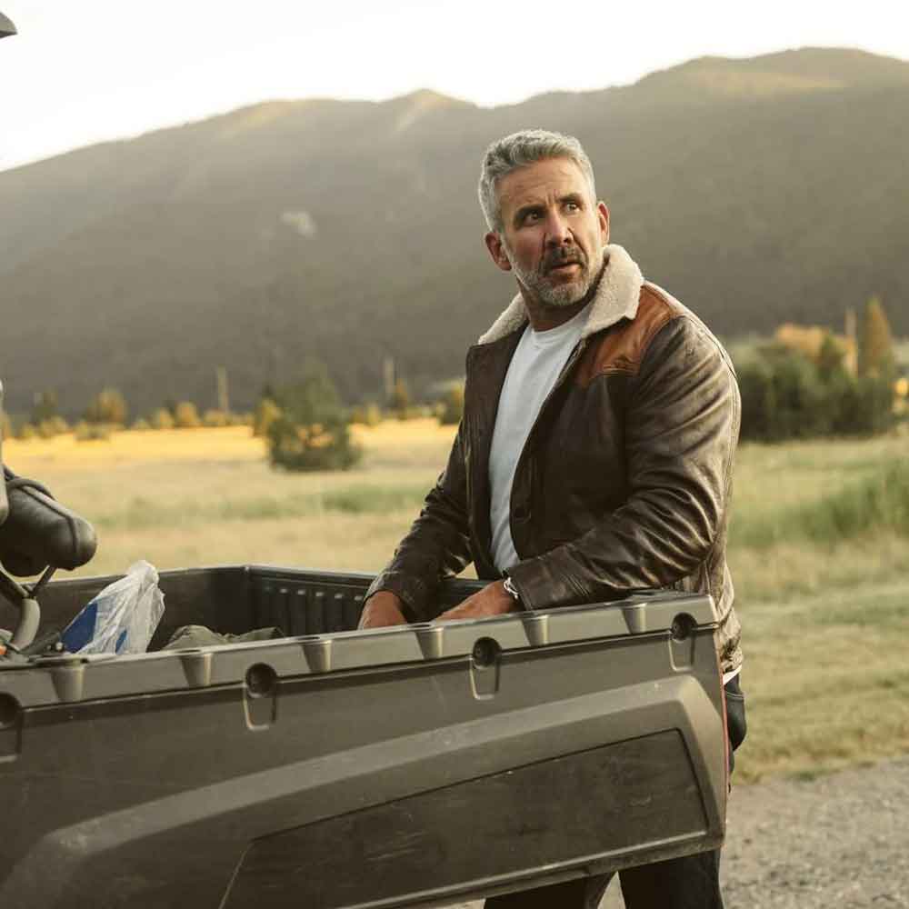 A man with gray hair and a beard wearing a brown leather jacket with a shearling collar, leaning on the tailgate of a dark utility truck in an outdoor field with mountains in the background.