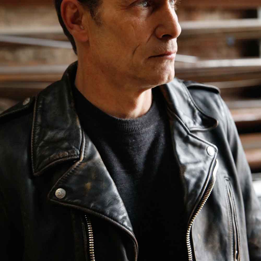 A close-up shot of a man wearing a black leather motorcycle jacket over a black crewneck shirt, set against a blurred background of wooden stairs.